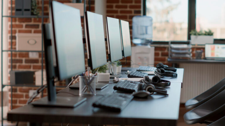 A row of desktop monitors, keyboards, and headsets in a workspace